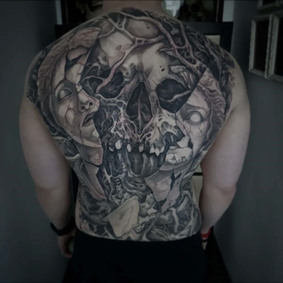 Espalda cubierta por un tatuaje negro y gris con rostros, calaveras y formas orgánicas.
