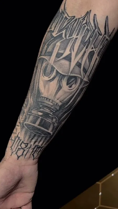 Tatuaje de una máscara y letras estilizadas en negro y gris en el antebrazo.